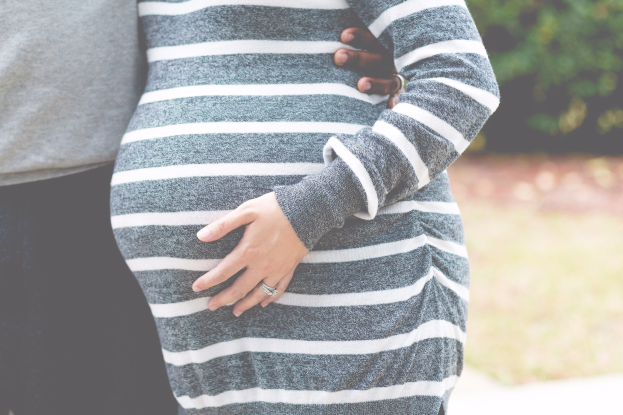 Schwangeres Paar, das Hand in Hand mit Händen auf dem Bauch steht, trägt Schwangerschaftskleidung und ist von Bäumen umgeben.
