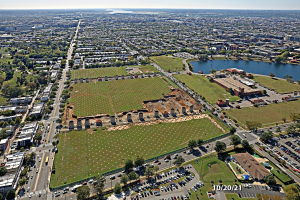 Luftaufnahme einer Stadt mit Gebäuden, Bäumen, Gras, Straßen mit Fahrzeugen, Wasser und Wolken, die einen großen zentralen Platz zeigt, der als Parkgebiet vorgeschlagen ist.