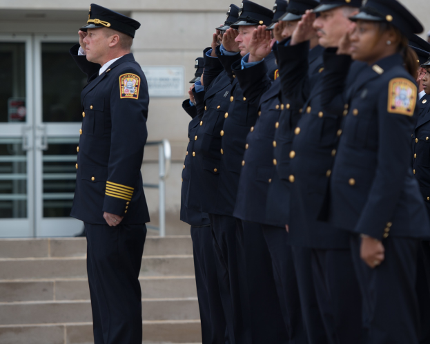 Eine Gruppe von Polizisten in Uniform und mit Mützen, die in Formation stehen und vor einem Gebäude mit Glastüren und Treppe salutieren.