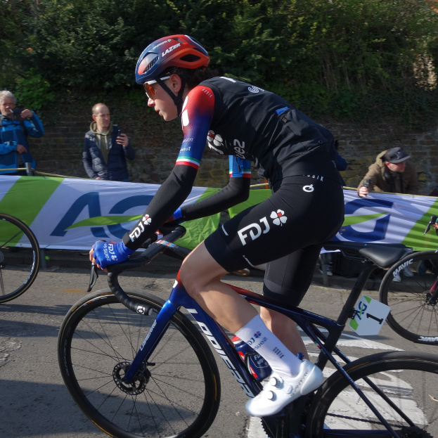 Professionelle Radrennfahrerin Katie Archibald trägt einen Helm, eine Schutzbrille, Handschuhe und Schuhe und fährt ein Fahrrad an einer Zuschauermenge vorbei, mit einem Banner, einer Wand, Bäumen und einem Gebäude im Hintergrund.