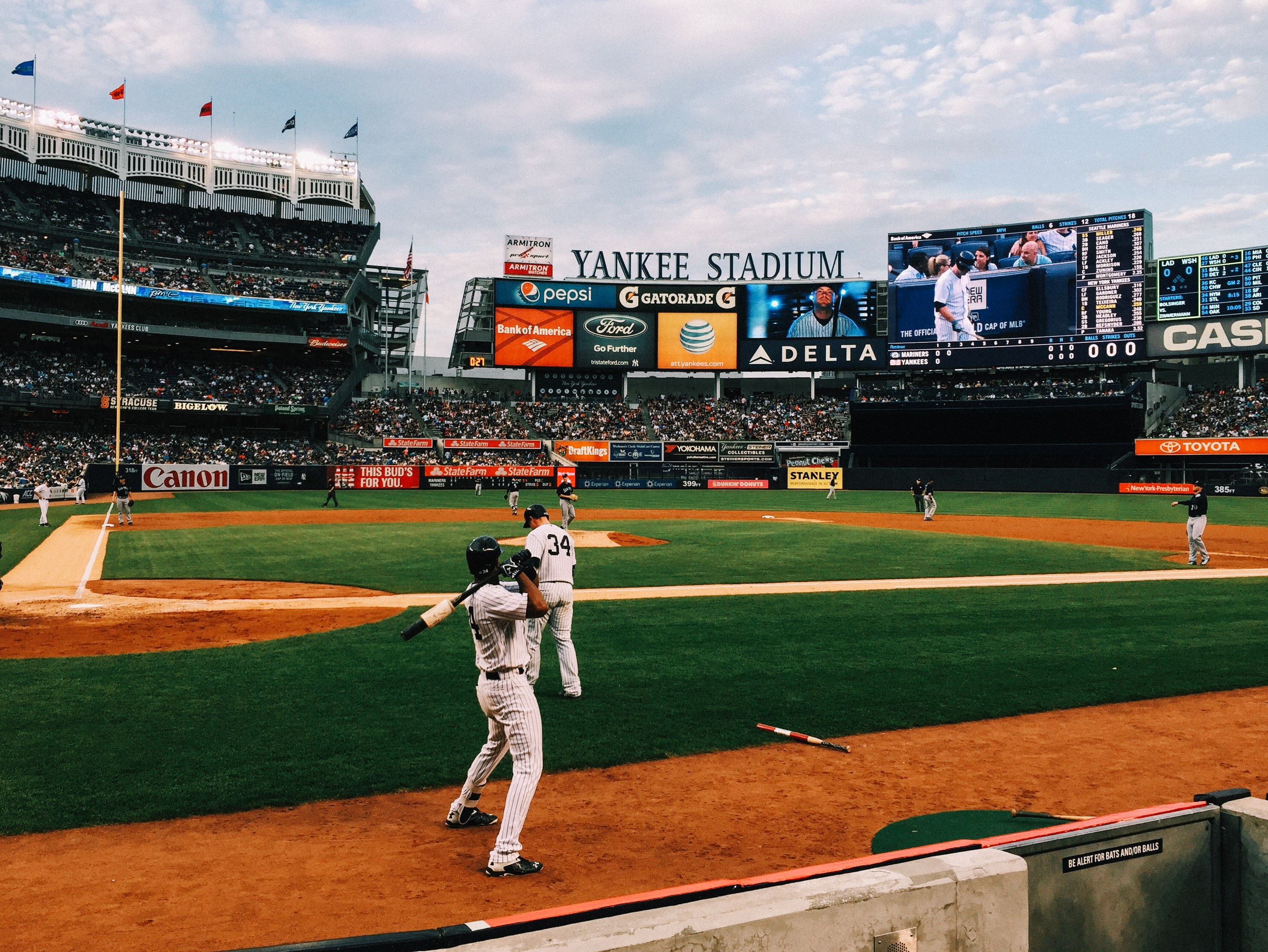 Baseballspiel im Yankee Stadium mit Spielern auf dem Feld und Zuschauern in den Rängen, umgeben von Stadionmerkmalen wie Zäunen, Flaggen, Schildern, einer Anzeigetafel und Deckenleuchten unter einem bewölktem Himmel.