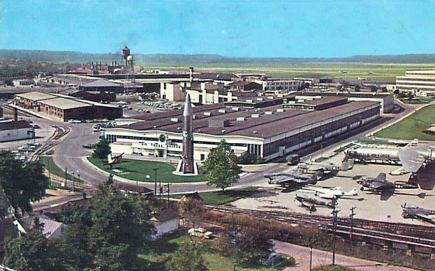 Alte Schwarz-Weiß-Flughafenszene mit geparkten Propellerflugzeugen, Terminalgebäuden, Bäumen, Straßen, Fahrzeugen, Versorgungsmasten, Gras, Bahnschienen, fernen Hügeln und einem klaren Himmel; Bildunterschrift unten.