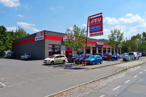 Ein Autohaus mit parkenden Fahrzeugen, umgeben von Gebäuden, Bäumen, Strommasten, Schildern und einer Straße, unter einem bewölkten Himmel.