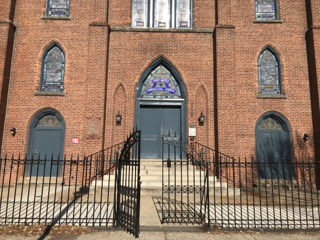 Ein großes Backsteingebäude mit einem schwarzen Tor davor, das Fenster, Türen, Treppen, Geländer und Lampen aufweist und als die First United Evangelical Church in Chicago identifiziert wird, mit einer Straße unten.