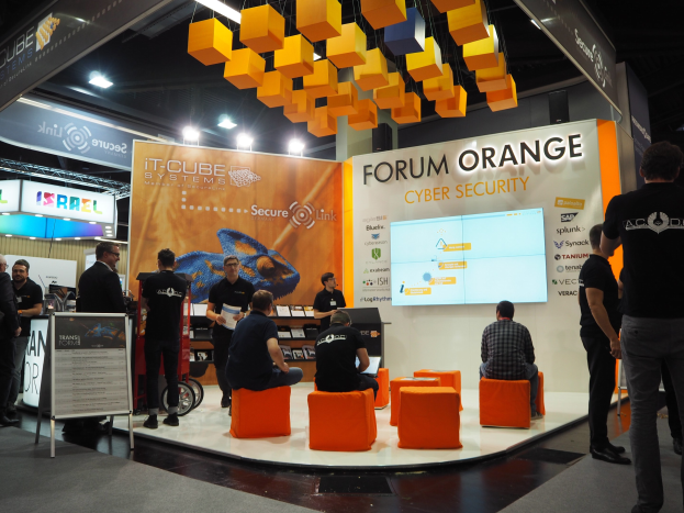 Eine Gruppe von Menschen an einem IT-Cube Systems Forum Orange Cyber Security Stand, mit sitzenden und stehenden Teilnehmern, einem Bildschirm, Regalen, Schildern, Deckenleuchten und einer Tafel mit Text.