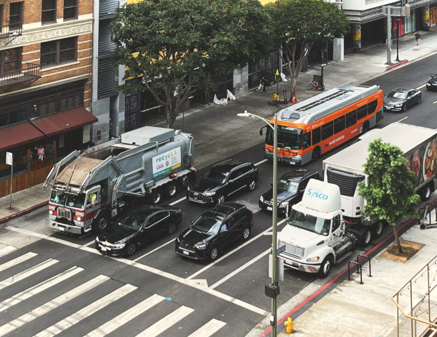 Eine belebte Stadtstraße mit Autos, Bussen, Lastwagen, Fußgängern, Bäumen, Laternen, Gebäuden, Geländern, Stufen und Schildern am Straßenrand.