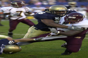 Zwei Fußballspieler in Helmen und Uniformen kämpfen auf dem Feld gegeneinander, während andere zuschauen.