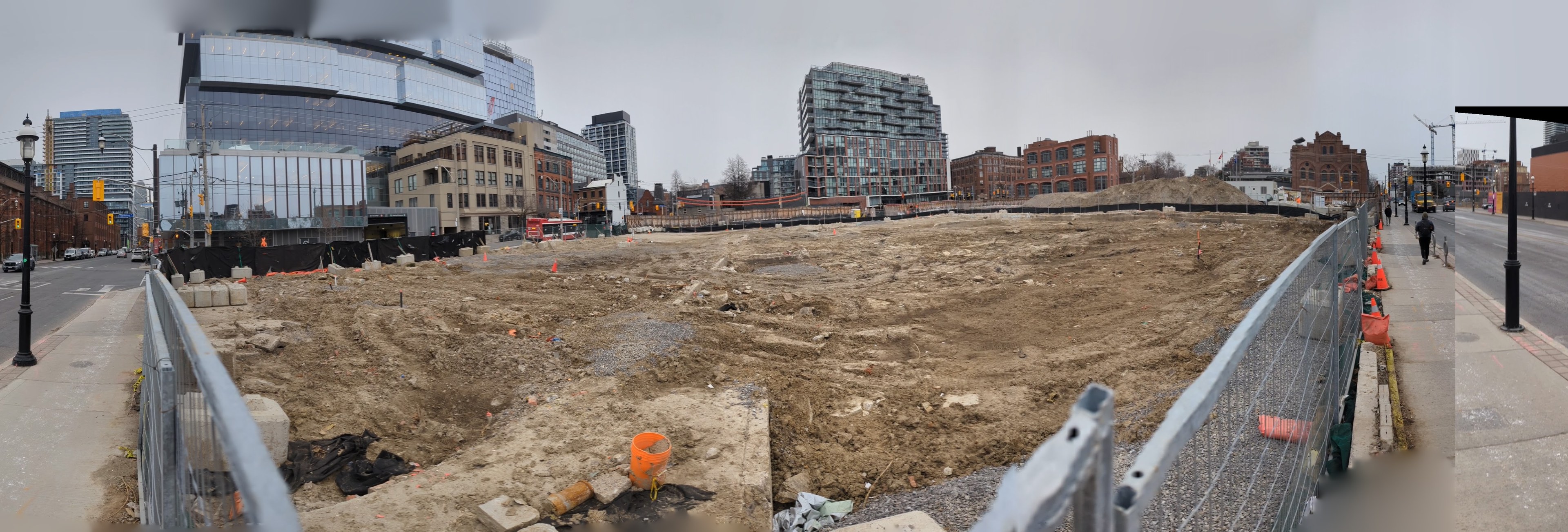 Großer Schutthaufen auf einer Baustelle umgeben von Gebäuden, Straßeninfrastruktur, Fahrzeugen, Fußgängern, Bäumen und einem klaren blauen Himmel.