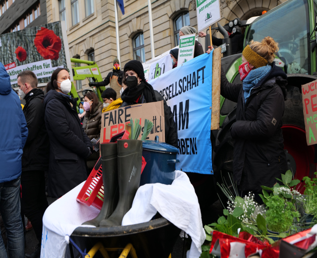 Eine Gruppe maskierter Menschen mit Protestschildern vor einem Lastwagen, mit einem Tisch voller Gegenstände, Topfpflanzen und einem flaggengeschmückten Gebäude im Hintergrund.