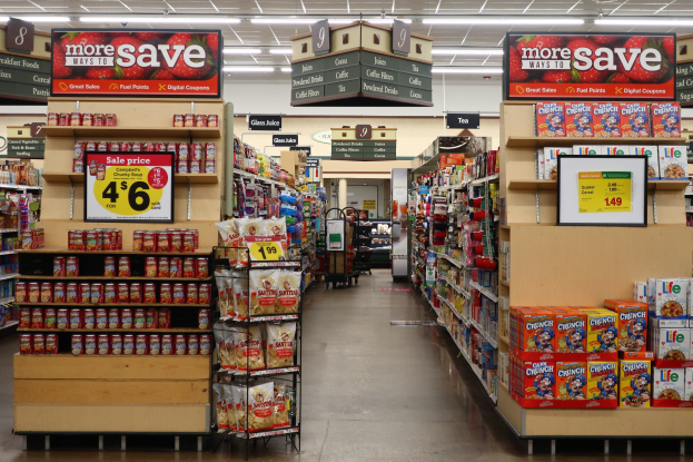 Ein Kiosk-Innenraum mit Regalen und Boden voller verschiedener Lebensmittel, Deckenbeleuchtung und Informationsschilder im Hintergrund, mit einer Person im Hintergrund sichtbar.