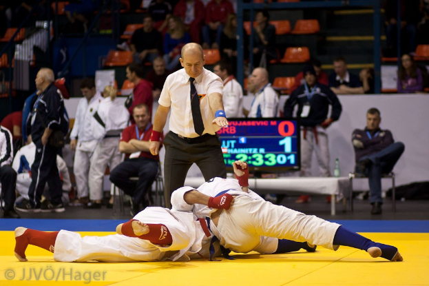 Zwei Judoka im Kampf mit einem Schiedsrichter in der Nähe, Zuschauern im Hintergrund und einer Anzeigetafel.