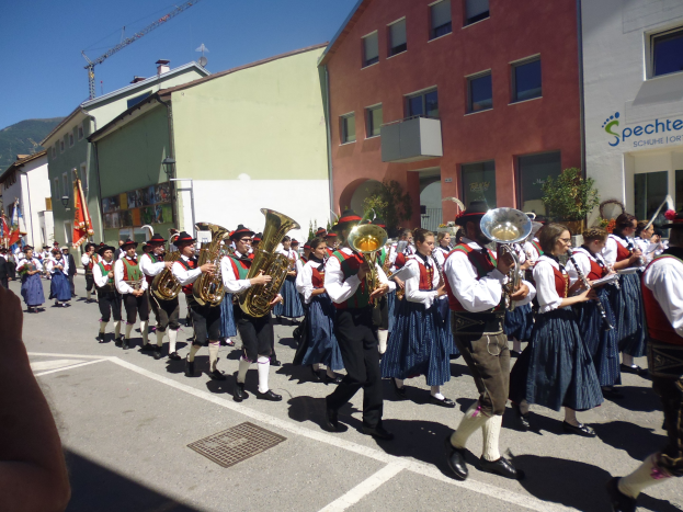 Eine Gruppe von Menschen in traditioneller bayrischer Tracht, die auf der Straße musizieren und dabei durch eine Straße mit Gebäuden gehen, einige halten Fahnen, im Hintergrund ein Hügel und ein blauer Himmel.
