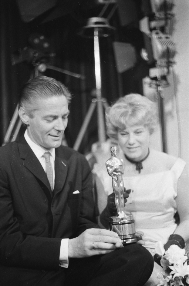 Ein Schwarz-Wei├č-Bild eines formell gekleideten Mannes, der eine Oscar-Statue und eine Frau mit einem Blumenstrau├čus in den Händen hält, die neben ihm sitzt, mit beleuchtetem Hintergrund.