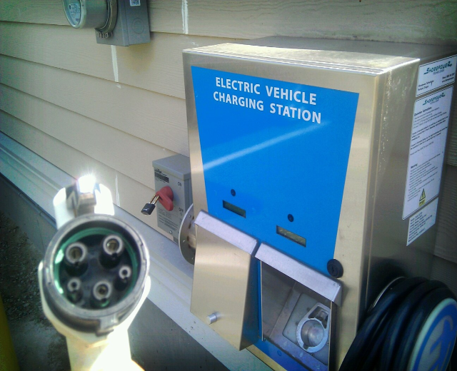 Elektrofahrzeug-Ladestation an der Seite eines Gebäudes mit angeschlossenem Ladekabel und zusätzlichen wandmontierten Gegenständen im Hintergrund.