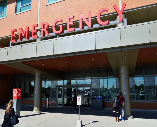 Gruppe von Menschen, die vor einem Notaufnahmeschalter mit Glaswänden, Säulen und Schildern gehen, einschließlich eines Mülleimers.