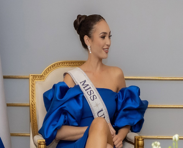 Frau in einem blauen Kleid mit einer "Miss Universe 2019" Schärpe, die lächelt, während sie auf einem Stuhl sitzt, umgeben von Blumen.