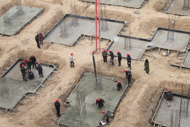 Gruppe von Bauarbeitern mit Helmen auf einer Baustelle, einige halten Werkzeuge, während andere Beton gießen; Eisenstangen und verstreute Materialien sichtbar.
