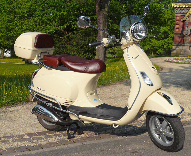 Ein Vespa GTS 300 Super ABS Roller auf einer Straße, umgeben von Bäumen, blühenden Pflanzen und einem Gebäude im Hintergrund.