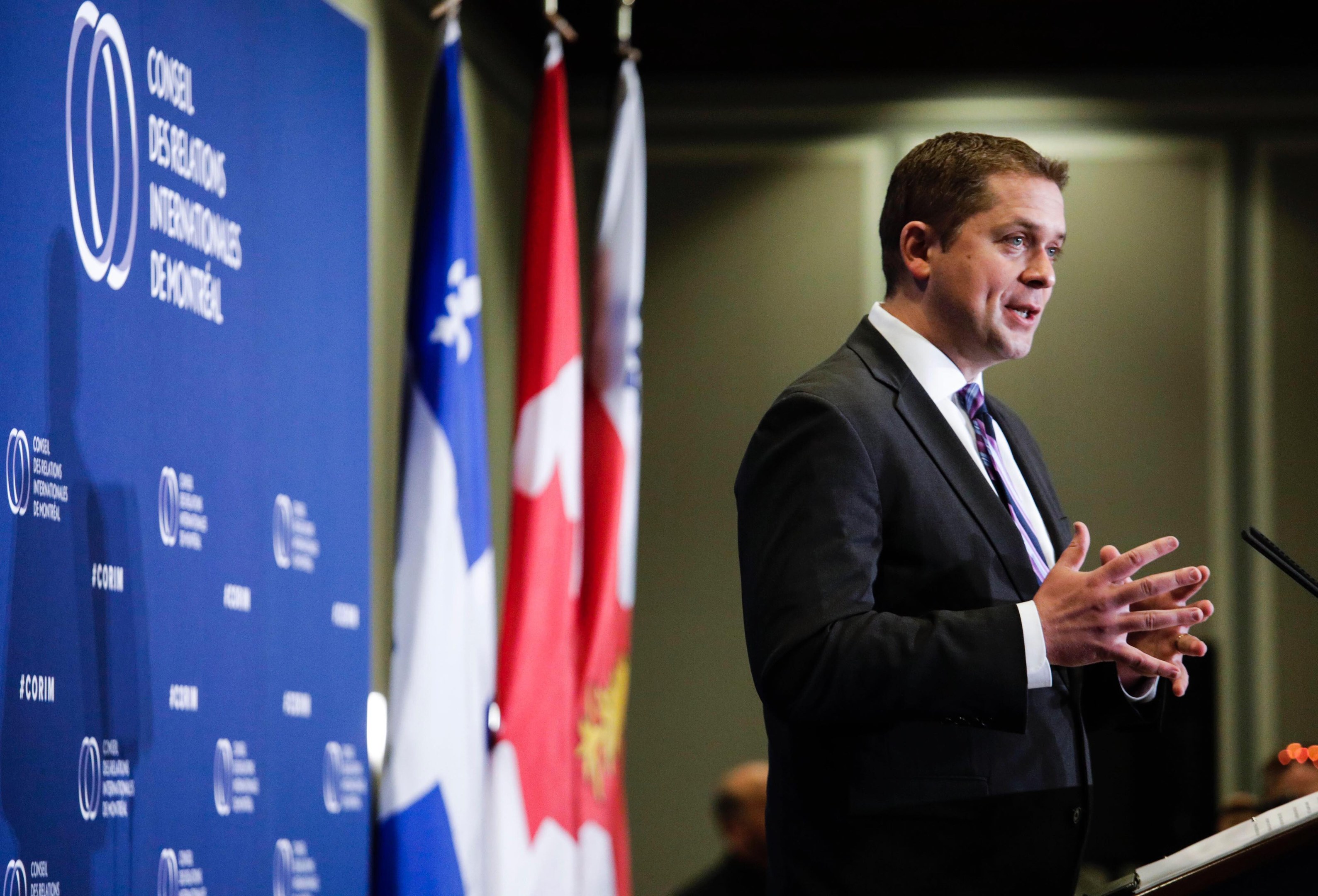 Kanadischer Premierminister an einem Rednerpult stehend, umgeben von Flaggen und einem Banner, mit Menschen im Hintergrund.