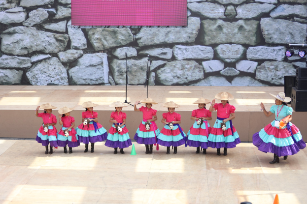 Gruppe junger Mädchen in bunten Kleidern und Hüten auf einer Bühne mit Verkehrskegeln, Mikrofonen, Lautsprechern, Lichtern und einem Hintergrundbild tanzen, während das Publikum unten zuschaut.