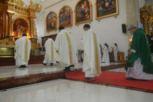 Eine Gruppe von Priestern geht einen roten Teppich in einer Kirche entlang, mit Menschen, die auf dem Boden vor ihnen und auf der rechten Seite stehen, und Fotorahmen an der Wand im Hintergrund.