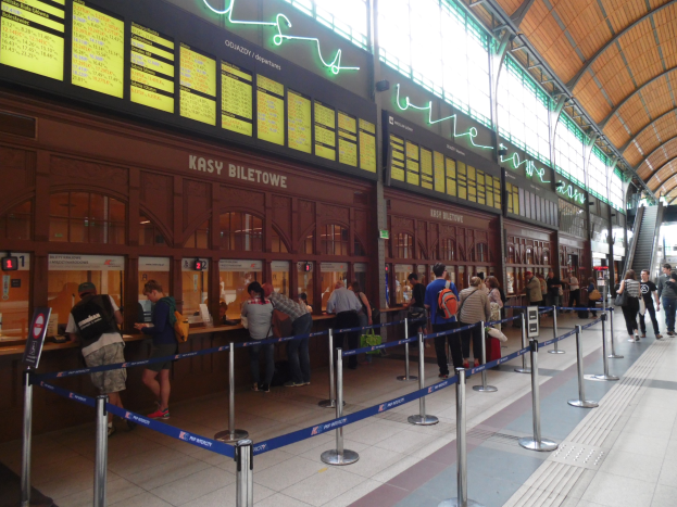 Menschen in einer Warteschlange an einem Bahnhofsschalter mit Deckenleuchten, Informationsanzeigen und ribbonverzierten Pfosten.