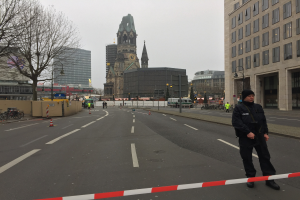 Ein Polizeibeamter in schwarzer Uniform und Mütze steht auf einer Straße in Berlin umgeben von Verkehrskegeln, Fahrrädern, Laternenpfählen, Bäumen und Gebäuden, im Hintergrund ist ein Kirchturm und ein klarer blauer Himmel zu sehen.