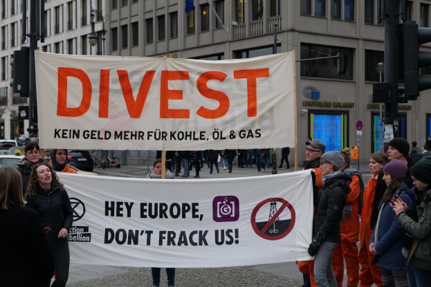 Menschen mit einem Banner mit der Aufschrift "Divest Hey Europa, Don't Frack Us" auf einer Straße mit Fahrzeugen, Gebäuden und Bäumen im Hintergrund.