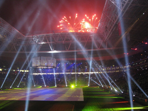 Olympisches Stadion in London bei Nacht, erleuchtet von hellen Lichtern und Feuerwerken, mit Menschen drinnen und einem großen Bildschirm im Vordergrund.