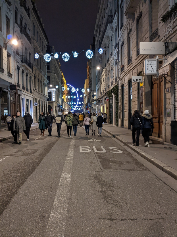 Menschen, die nachts durch eine Straße in Barcelona gehen, beleuchtet von festlichen Weihnachtslichtern, mit Gebäuden auf beiden Seiten und dem Himmel im Hintergrund.