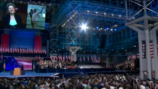 Eine große Menschenmenge sitzt vor einer Bühne mit einem Podium, Mikrofon, Flaggen, einem Bildschirm und Beleuchtungsausrüstung und wartet auf eine Präsidentschaftsdebatte.