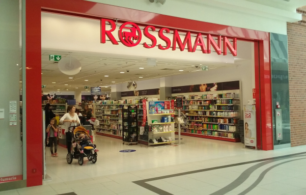 Ein Rossmann-Laden in einem Einkaufszentrum mit Menschen, einer Person mit einem Kinderwagen und Regalen mit Produkten unter Deckenleuchten und Schildern.