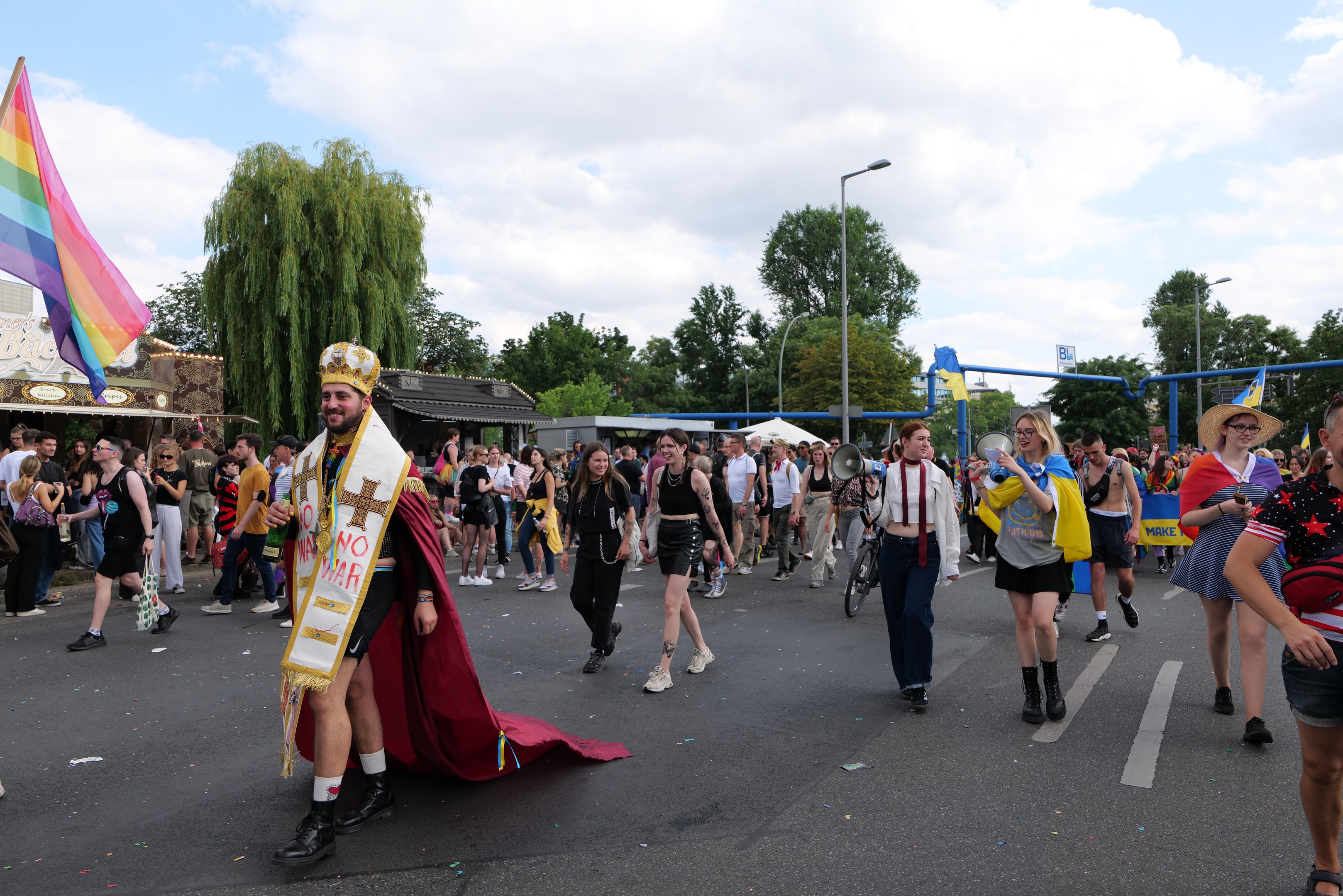 Eine Gruppe von Menschen marschiert bei der Gay Pride Parade 2018 mit einer Regenbogenfahne und Musikinstrumenten, mit Laternenmasten, Bäumen und Hütten im Hintergrund.