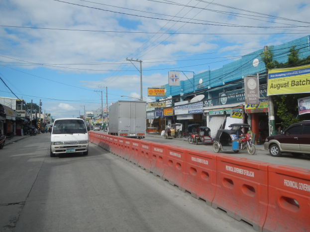 Eine belebte Stadtstraße mit starkem Verkehr, einschließlich Autos, einer Mitteltrennwand, Gebäuden mit Schildern, Strommasten mit Überlandleitungen, verstreuten Bäumen und einem bewölkten Himmel.