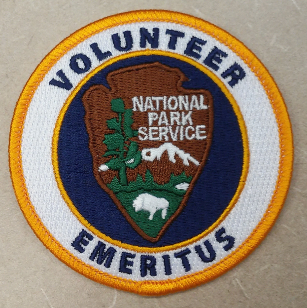 Runder Aufnäher mit blauem Rand und weißem Hintergrund, der den schwarzen Text 'Volunteer National Park Service Emeritus' in einem gelben Kreis in der Mitte zeigt.