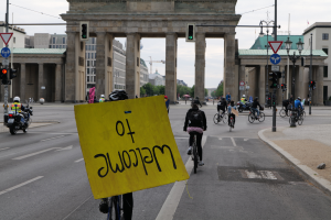 Eine Gruppe von Menschen auf Fahrrädern fährt eine Straße vor dem Brandenburgertor in Berlin, Deutschland, entlang, trägt Helme und hält ein gelbes Schild hoch, mit Gebäuden, Bäumen und einem klaren blauen Himmel im Hintergrund.