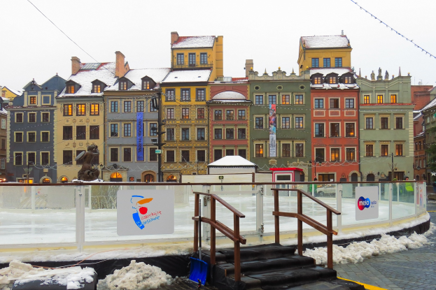 Eislaufbahn in einem Stadtplatz umgeben von Gebäuden, Treppen, Geländern, Informationsschildern, schneebedecktem Boden, Laternen, Bäumen und Himmel.