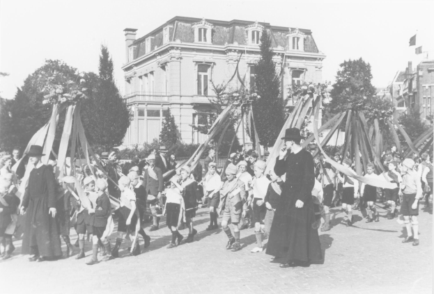 Eine Gruppe von Menschen in Schwarz-Weiß auf einer Straße mit Fahnen, einem Maibaum und Gebäuden im Hintergrund.