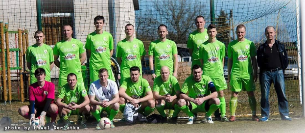Eine Gruppe von Männern in grünen T-Shirts steht auf einem Fußballfeld in der Nähe eines Torpfostens, mit einem Ball davor, Bäume und Gebäude im Hintergrund.