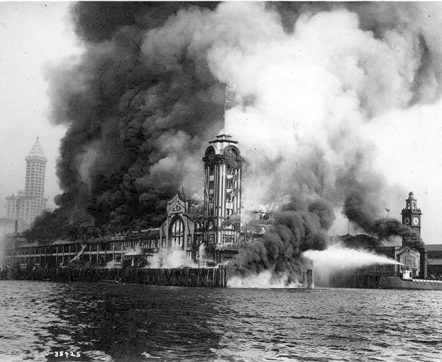 Schwarzes und weißes Foto eines großen Gebäudes, das von Flammen eingehüllt ist, mit Rauch, der herausquillt, einem Gewässer im Vordergrund und anderen Gebäuden und dem Himmel im Hintergrund.