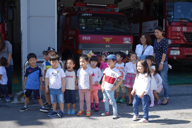 Eine Gruppe von Kindern vor einem Feuerwehrauto auf einem Feuerwehrhof, einige tragen Mützen, mit zusätzlichen Feuerwehrautos im Hintergrund.