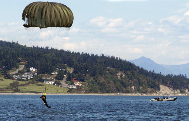 Eine Person gleitet mit einem Gleitschirm über einen See mit einem Boot auf der rechten Seite, Bäume, Gebäude, Hügel und einen klaren blauen Himmel im Hintergrund.