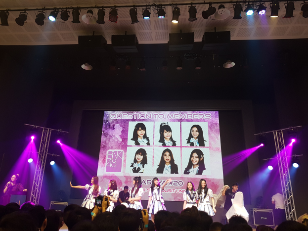 Eine Bühnenperformance mit zwei Personen: einer, die ein Mikrofon hält und spricht, und einer, die ein Instrument spielt, mit hellen Lichtern und Trägern darüber. Unten eine Menge mit Kameras, während ein großer Bildschirm im Hintergrund Bilder der K-Pop-Gruppe BK48 anzeigt.