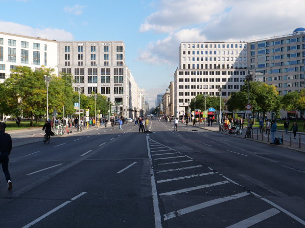 Eine belebte Stadtstraße in Berlin, Deutschland, mit Fußgängern und Radfahrern auf der Straße, hohen Gebäuden mit Fenstern, Bäumen, Laternen und Schildern, unter einem bewölkten Himmel.