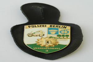 Ein schildförmiges Polizeisiegel mit der Aufschrift "Polizei Berlin" in schwarzer fetter Schrift auf einem blauen Hintergrund mit einem weißen Stern und Rahmen, das auf einer Oberfläche liegt.