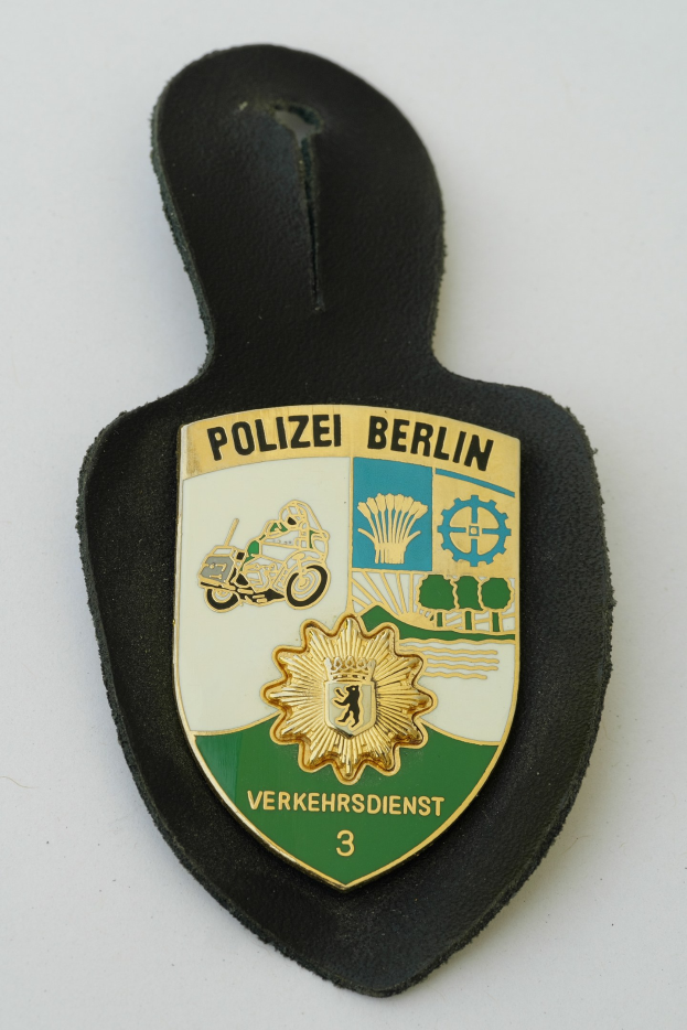 Ein schildförmiges Polizeisiegel mit der Aufschrift "Polizei Berlin" in schwarzer fetter Schrift auf einem blauen Hintergrund mit einem weißen Stern und Rahmen, das auf einer Oberfläche liegt.