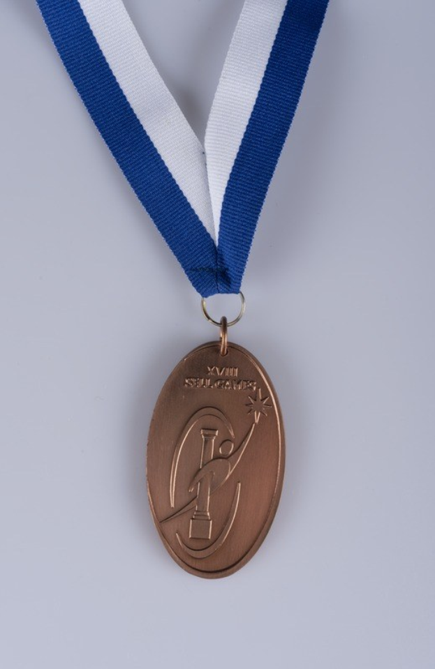 Goldene Medaille mit blau-weißem Band, eingravierter Schrift, auf weißem Hintergrund.
