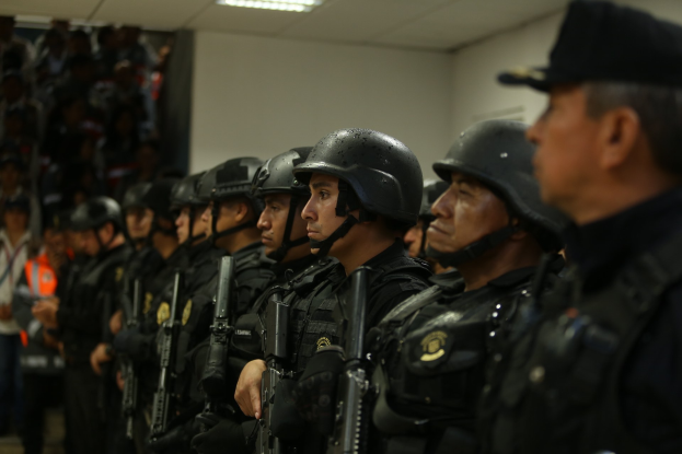 Eine Gruppe von Polizisten in Helmen und taktischer Ausrüstung steht in Formation, hält Gewehre und hat eine Wand und Deckenleuchten im Hintergrund.