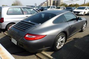 Ein silberner Porsche 911 auf einem Parkplatz mit anderen Autos, Gebäuden, Bäumen, Pfählen, Drähten und einem klaren blauen Himmel.