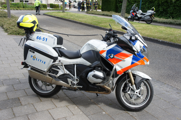Ein Polizei-Motorrad am Straßenrand mit einer Gruppe von Menschen in bunten Kleidern und Helmen, umgeben von Bäumen, Pflanzen und Gebäuden im Hintergrund.